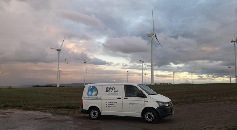 Weißer Transporter vor Windkraftanlagen unter bewölktem Himmel.