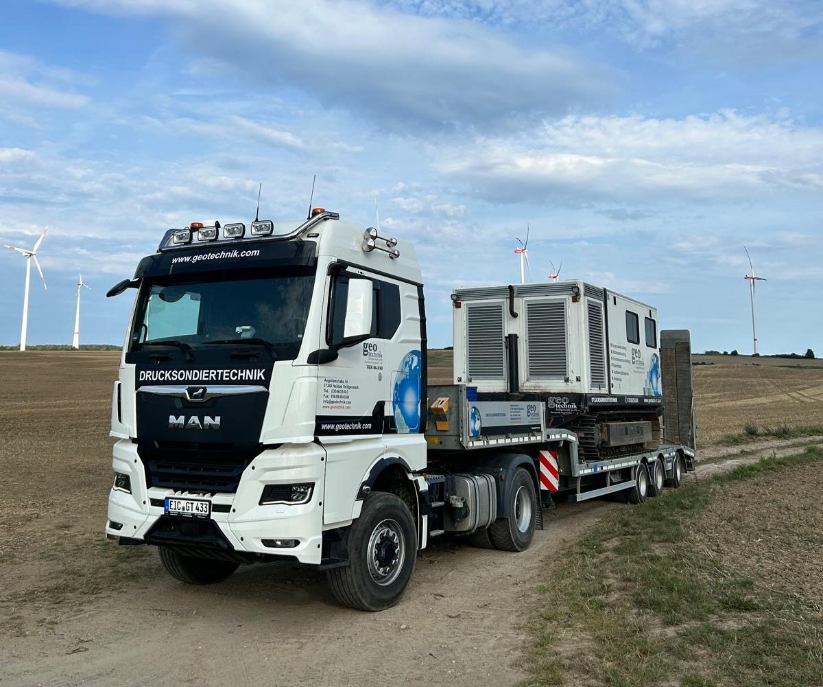 Lkw für Drucksondiertechnik mit einem Anhänger, der eine mobile Einheit transportiert, vor einer Landschaft mit Windrädern
