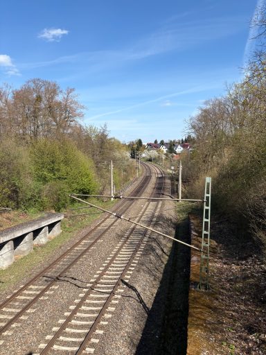 Eisenbahngleise mit Bäumen und einem Dorf im Hintergrund unter klarem Himmel.