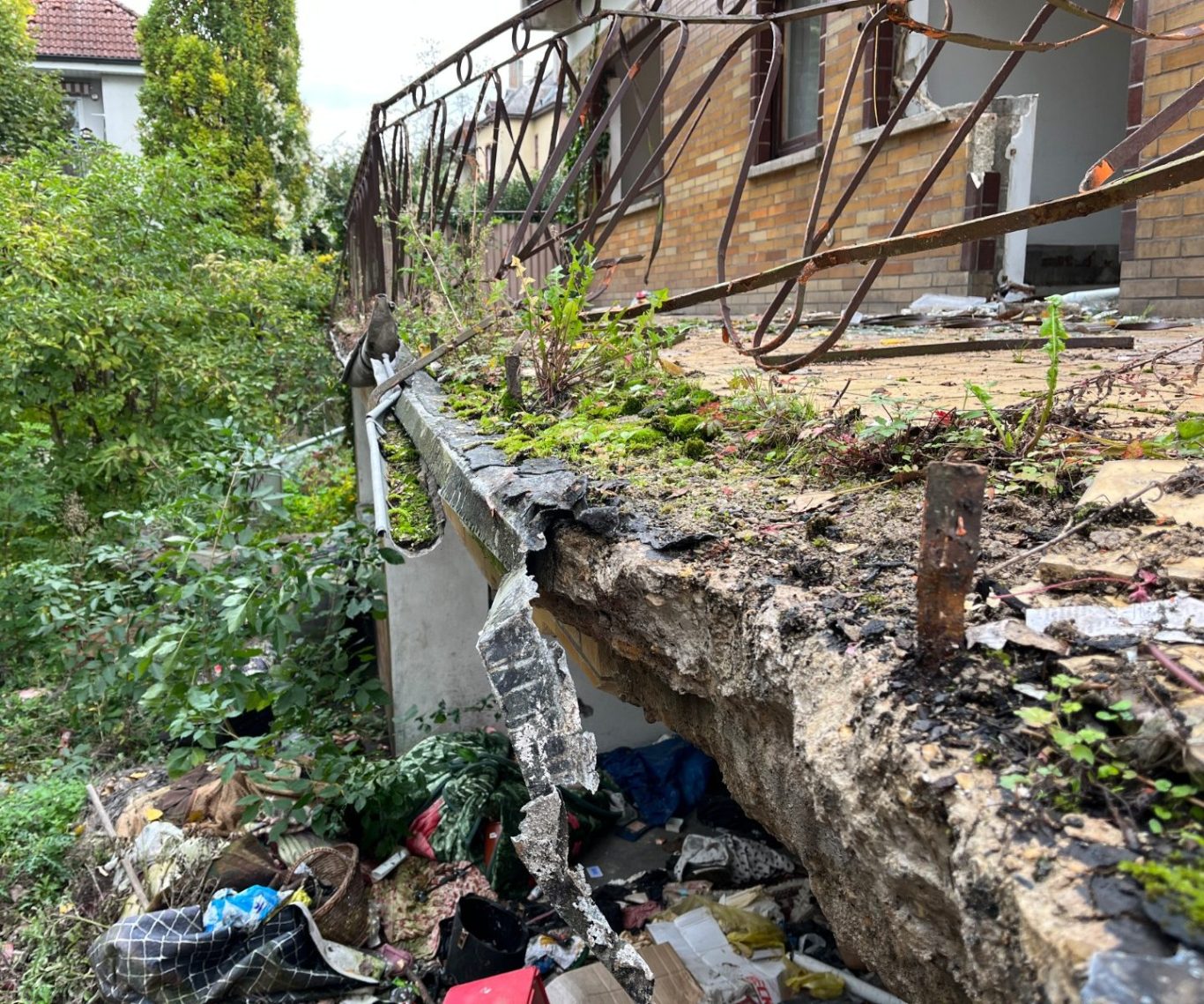 Ein vernachlässigtes, verschmutztes Gebäude mit beschädigtem Fundament, das von Müll und dichter Vegetation umgeben ist.