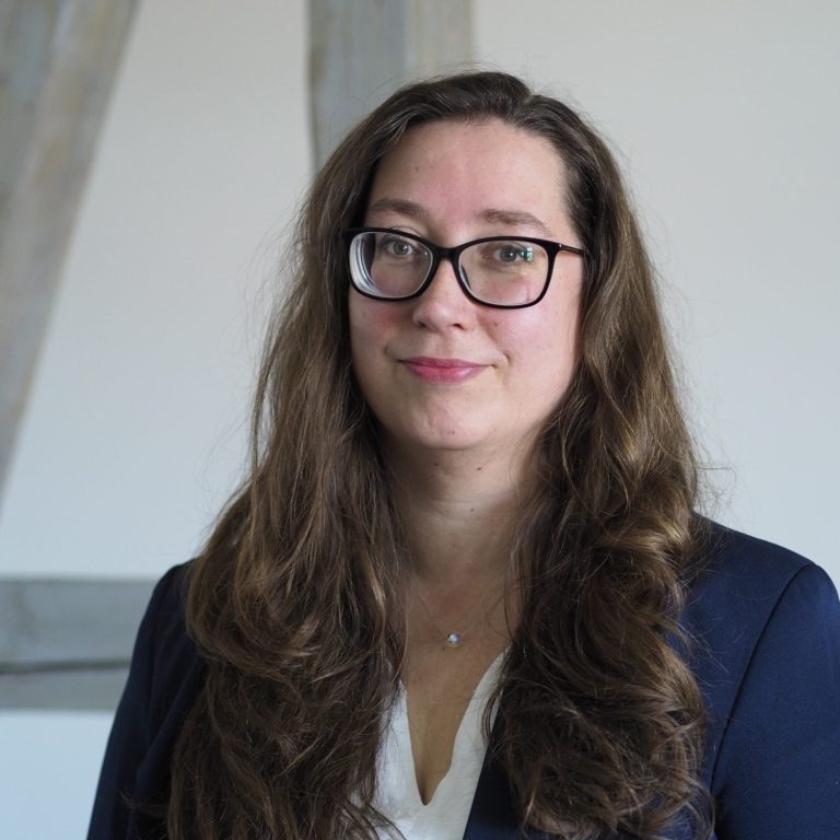 Karola Raub mit langen, lockigen Haaren, Brille und blauen Augen, trägt ein dunkelblaues Outfit.